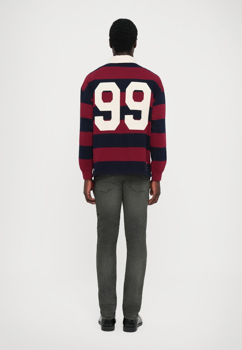 Navy en bordeaux gestreept rugbyshirt met het witte nummer "99" op de achterkant; gecombineerd met grijze jeans en zwarte schoenen, gezien vanuit de achterkant.