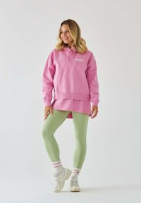 Przytulny różowy hoodie w oversize z kieszenią z przodu i logo, w połączeniu z jasnoróżowymi legginsami oraz białymi sneakersami z różowymi akcentami i paskowanymi skarpetkami.