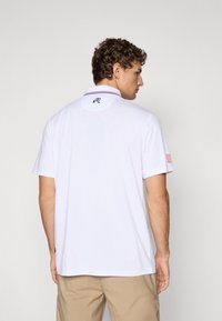 White polo shirt with short sleeves, red and blue accents on collar, shoulder patch featuring American flag, and embroidered logo on back.