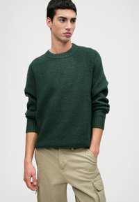 Mørkegrøn strikket sweater med ribbet tekstur, rund halsudskæring og lange ærmer, parret med lyse beige cargo bukser.