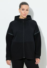 Sudadera negra con cremallera, con una sección inferior texturizada, mangas raglán, capucha ajustable con cordones y detalles reflectantes en la parte superior del pecho.