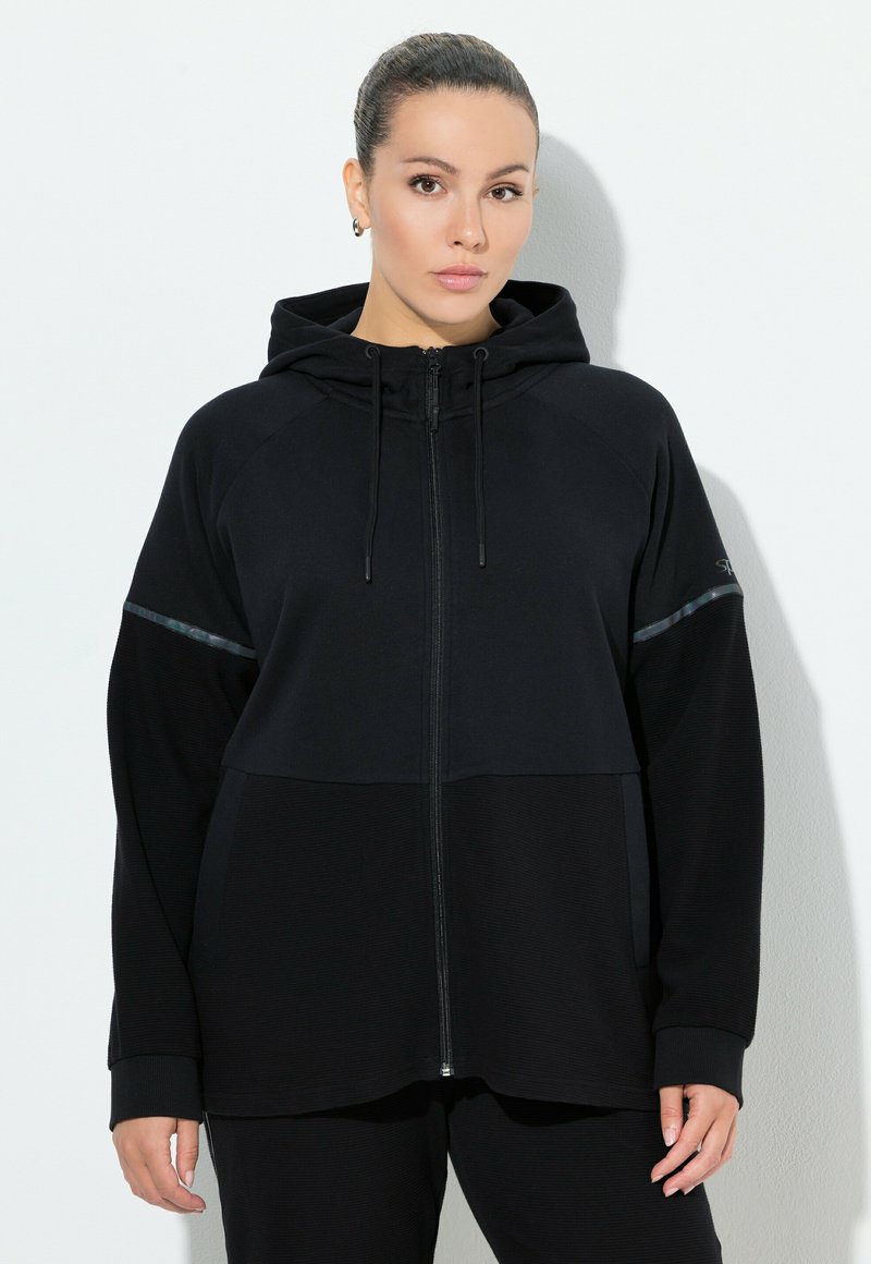 Sudadera negra con cremallera, con una sección inferior texturizada, mangas raglán, capucha ajustable con cordones y detalles reflectantes en la parte superior del pecho.