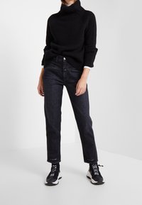 Jersey de cuello alto negro acanalado, vaqueros oscuros y zapatillas negras con detalles en blanco, posando sobre un fondo neutro.