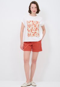 Weißes Kurzarmoberteil mit einem floralen Muster in Orange und Creme, kombiniert mit rostfarbenen Shorts und hellen Slippern.