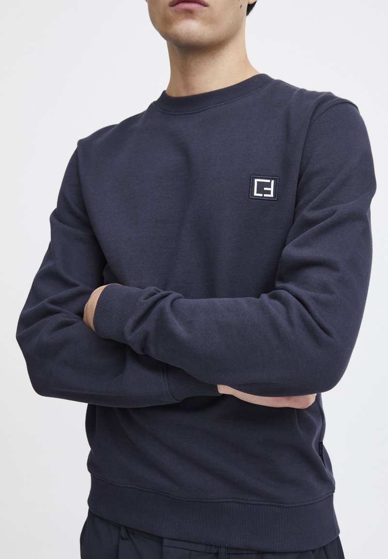 Man wearing dark navy crewneck sweatshirt with small white and navy patch logo on left chest, arms crossed against white background.
