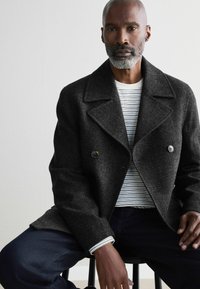 Abrigo de lana gris de doble botonadura con grandes solapas, combinado con una camisa blanca y azul marino de rayas. Modelo sentado, con jeans azul oscuro.