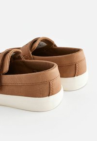 Bruna tygskor med en slät yta, som har en vit gummisula, sydda detaljer och en öppen häldesign för enkel påtagning.