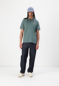 Zelený polo tričko s malým logom, tmavé denimové džínsy a svetlomodrá čiapka. Biele masívne tenisky dotvárajú outfit.