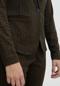 Gros plan d'une personne portant un costume en chevrons marron et noir avec une veste boutonnée et les mains détendues le long du corps.