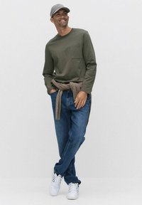 Långärmad olivgrön skjorta med bröstficka, kombinerad med blå jeans och vita sneakers. En brun stickad artikel är knuten runt midjan.