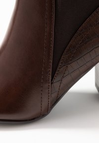 Botín de piel marrón hasta el tobillo con un patrón texturizado de cocodrilo en el tacón y paneles laterales elásticos, con detalle de costura visible.