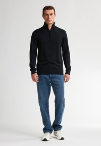 Schwarzer Reißverschluss-Pullover aus Strickstoff, kombiniert mit blauen Jeans und weißen Sneakern. Der Pullover hat einen hohen Kragen und enge Bündchen.