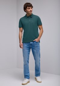 Mörkgrön polo-shirt med krage, parat med ljusblå jeans. Modellen står mot en enfärgad bakgrund, händerna i fickorna, avslappnad passform.