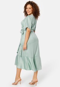 Een mintgroene satijnen midi-jurk met korte mouwen, een gestrikte taille en een uitlopende zoom. G gedragen met naakte, gestrikte hoge hakken.