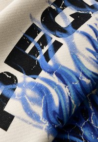 Tessuto di cotone con una base crema. Presenta testo grafico blu e nero con motivi a onda, finitura testurizzata e dettagli invecchiati.