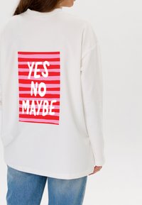 Witte sweatshirt met een losse pasvorm, met een achterafdruk: rood en roze gestreepte achtergrond met de tekst "JA NEE MISSCHIEN" in dikke witte letters.