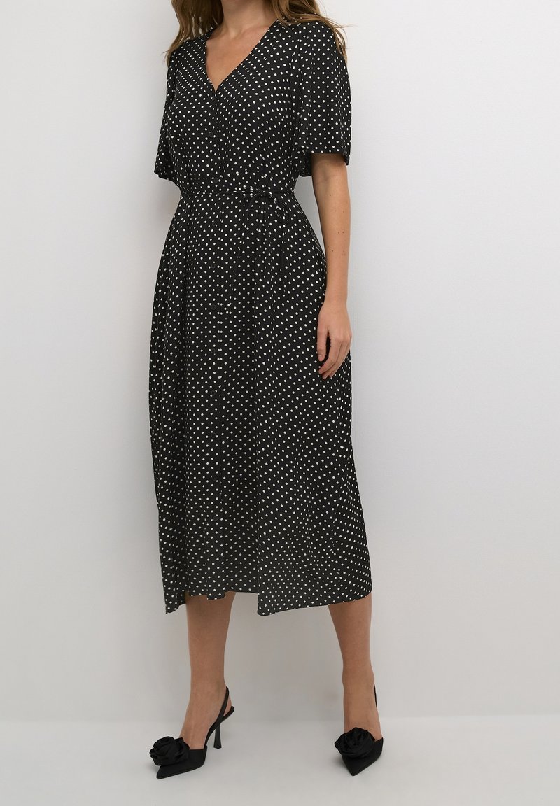 Femme portant une robe midi portefeuille noire à pois blancs avec manches courtes, associée à des talons noirs ornés de décorations florales.
