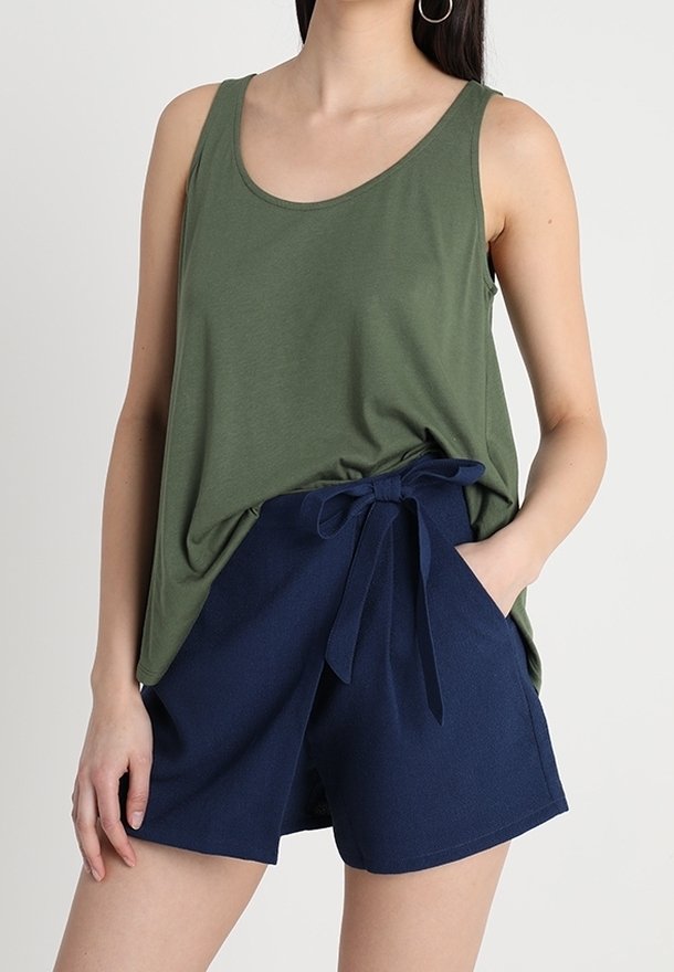 Woman wearing olive green sleeveless top and navy blue high-waist shorts with fabric belt tied in a bow, one hand in pocket.