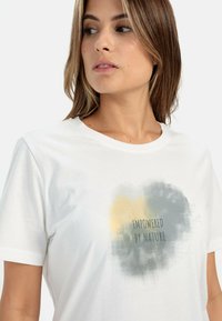 Hvid bomuld t-shirt med rund halsudskæring, korte ærmer og et tekstureret grafisk print med teksten "Empowered by Nature" i grå og gul.