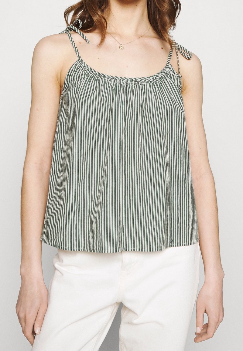 Femme portant un haut sans manches rayé verticalement gris et blanc avec des bretelles à nouer et un pantalon blanc, raccourci au niveau du cou et des hanches.