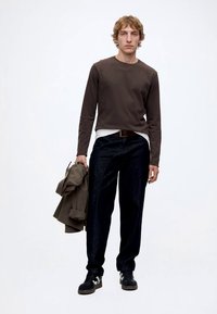 Junger Mann mit lockigem Haar, der ein dunkles Langarmshirt, schwarze Hose und Turnschuhe trägt und eine gefaltete Jacke in der linken Hand hält.