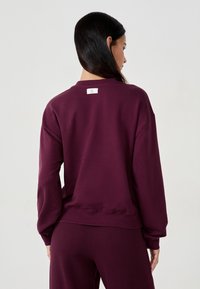 Felpa bordeaux con collo a giro e spalle cadenti, realizzata in tessuto morbido. Presenta un piccolo etichetta del marchio sul collo sul retro.