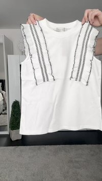 Top bianco senza maniche con bordo arricciato nero e cuciture a zigzag sulle spalle. Tessuto liscio, scollatura a girocollo e vestibilità morbida.