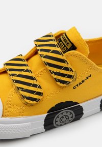 Gule lerrets-sneakers med svarte og gule stripete borrelåsremmer, gummisåle og et tegneseriehjuldesign på tåen.