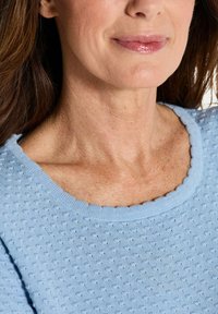 Vrouwelijke onder gezicht en hals met lichte make-up, gedragen een textuur in lichtblauwe top met een gegolfde halslijn.
