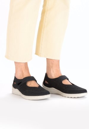 Black, perforated Mary Jane shoes with a strap, light gray sole, and cutouts on the sides, worn with light yellow cropped jeans.