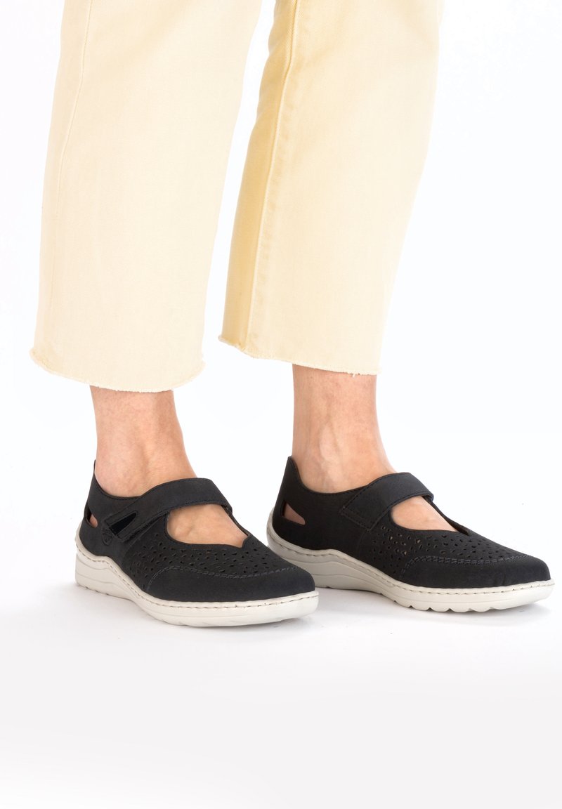 Schwarze, durchbrochene Mary Jane-Schuhe mit einem Riemen, hellgrauer Sohle und Ausschnitten an den Seiten, getragen mit hellgelben, kurzen Jeans.