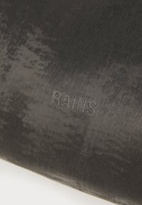Svart vanntett materiale med en teksturert overflate. Ordet "RAINS" er preget, noe som fremhever merket med et subtilt design.