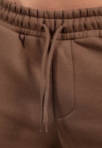 Brune sweatpants har en ribbet talje, snøre og sidelomme, lavet af et blødt, tekstureret stof med en glat overflade.