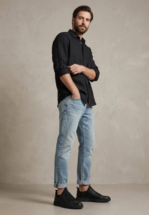 Man met baard die een zwart overhemd met opgerolde mouwen, lichtblauwe opgerolde jeans en zwarte sneakers draagt, staand tegen een effen muur.
