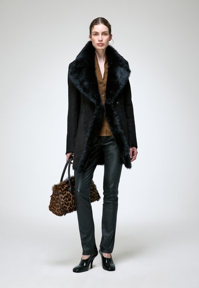 Femme portant un manteau noir bordé de fourrure, chemise marron, pantalon en cuir noir, talons noirs, tenant un sac à main en fourrure à motif léopard, debout sur un fond neutre.