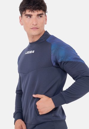 Muž nosící tmavě modrou mikinu s geometrickými modrými vzory na ramenou a logem „LEGEA“ na hrudi, ruka v postranní kapse.