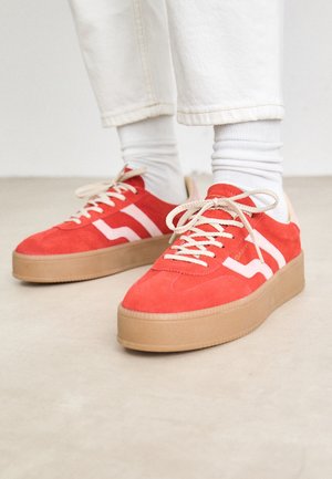 Personne portant un pantalon blanc et des chaussettes avec des baskets à plateforme en daim rouge, lacets et semelles beiges, sur un sol clair.