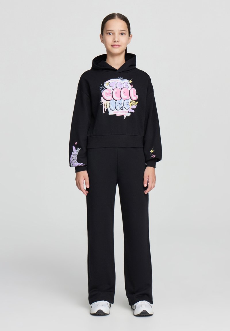 Felpa nera con stampa grafica colorata, polsini a costine e pantaloni della tuta neri abbinati. Sneakers bianche completano l'outfit.