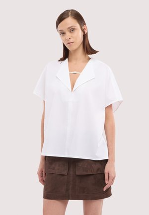 Frau mit glatten braunen Haaren trägt eine lockere weiße Bluse mit kurzen Ärmeln und tiefem Kragen sowie einen dunkelbraunen Rock mit großen Taschen.