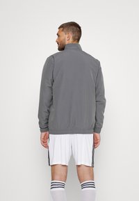 Grau Windbreaker-Jacke, elastische Bündchen, elastischer Saum, glatte Textur, minimalistisches Design, getragen über weißen Sportshorts und Socken.