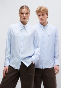 Ανοιχτό μπλε πουκάμισα με μεγάλο γιακά, υφασμάτινη υφή και αντιθετική λευκή επένδυση, σε συνδυασμό με σκούρα καφέ παντελόνια.