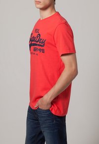 Camiseta de algodón roja con mangas cortas, que presenta un logo y texto impreso en azul marino "SuperDry" en la parte frontal. Corte casual, cuello redondo.