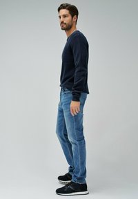 Granatowy, dziany sweter, jasnoniebieskie dżinsy, czarne trampki z białą podeszwą. Model stoi bokiem na neutralnym szarym tle.