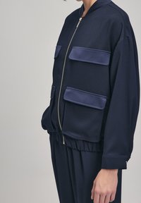 Marinblå bomberjacka tillverkad av ett slätt material, med två stora fackfickor, dragkedja fram och elastisk samling vid nederkant och ärmar.