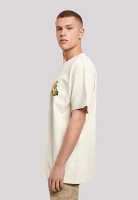 Cream-colored short-sleeve t-shirt with a loose fit. Features a graphic design on the left chest and a round neckline.