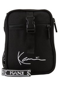 Borsa a tracolla nera in tessuto con zip, dotata di una tracolla regolabile e dettaglio bianco con firma sulla parte anteriore. Forma compatta e rettangolare.