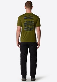 Olivově zelené tričko s krátkým rukávem, na kterém je černý grafický text a vzory na zadní straně, spárované s černými kalhotami a turistickými botami.