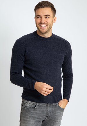 Glimlachende man met een baard, gekleed in een donkerblauwe gebreide trui en grijze jeans, staat tegen een plain witte achtergrond.
