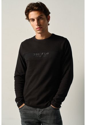 Joven con cabello corto y ondulado, vestido con una camisa negra de manga larga con texto japonés y vaqueros grises, de pie con las manos en los bolsillos.