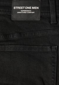 Primer plano de unos pantalones vaqueros negros que muestra una etiqueta que dice "STREET ONE MEN - Experience - Unmatched Comfort" cerca de la cintura.